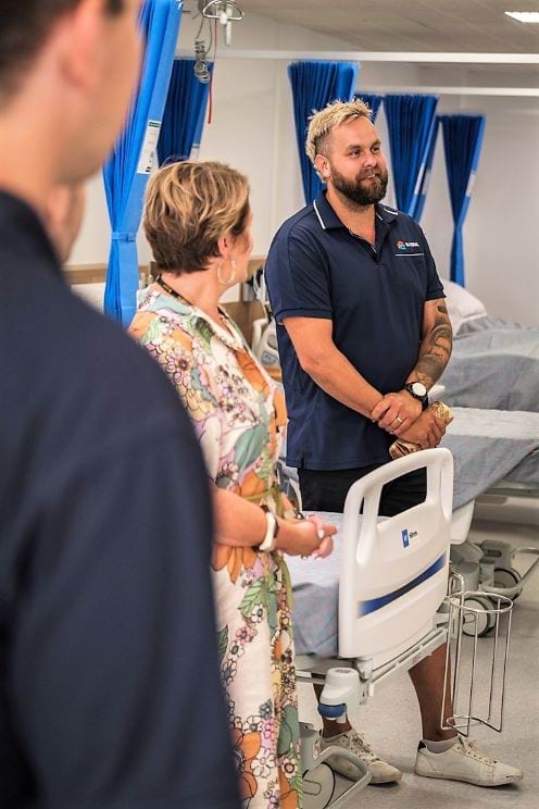 Man in Barpa polo t-shirt standing between hospital beds, looking off to camera right. A woman is looking away from camera and a partial silhouette of a man in extreme foreground is on left hand side.