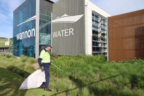 Man standing in garden with large white bag and litter picker. He is in front of a large building with 'Wannon Water' written on the side.