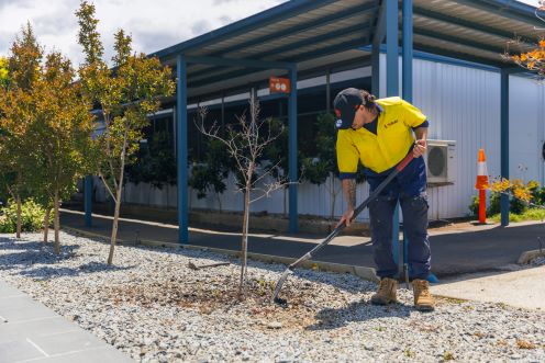 Young man in yellow high-vis top, leaning over with a shovel doing some landscaping. His face is obscured by the brim of his cap.