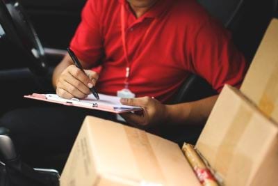 Photo of a delivery worker surrounded by packages and filling in a form on a clipboard